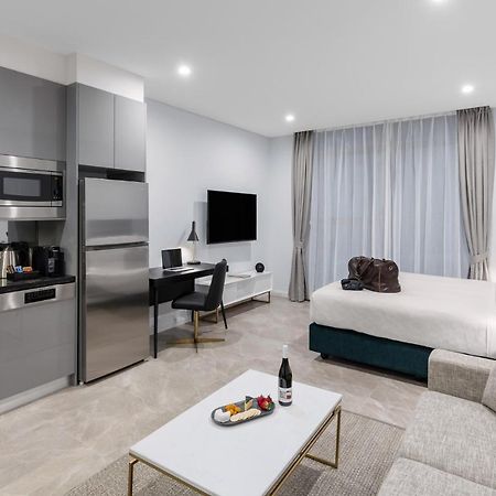 فندق أجنحة ميريتون سوسيكس ستريت سيدني المظهر الخارجي الصورة The photo shows a modern studio apartment. On one side, there is a kitchenette with sleek gray cabinets, a refrigerator, and built-in appliances. In the living area, there is a comfortable gray sofa, a white coffee table with a fruit platter and a bo
