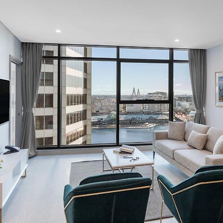 فندق أجنحة ميريتون سوسيكس ستريت سيدني المظهر الخارجي الصورة The photo shows a modern living room with large windows that offer a view of the cityscape and water. The interior features a light color palette with pale blue walls and soft lighting. There is a comfortable gray sofa and two green accent chairs, ar