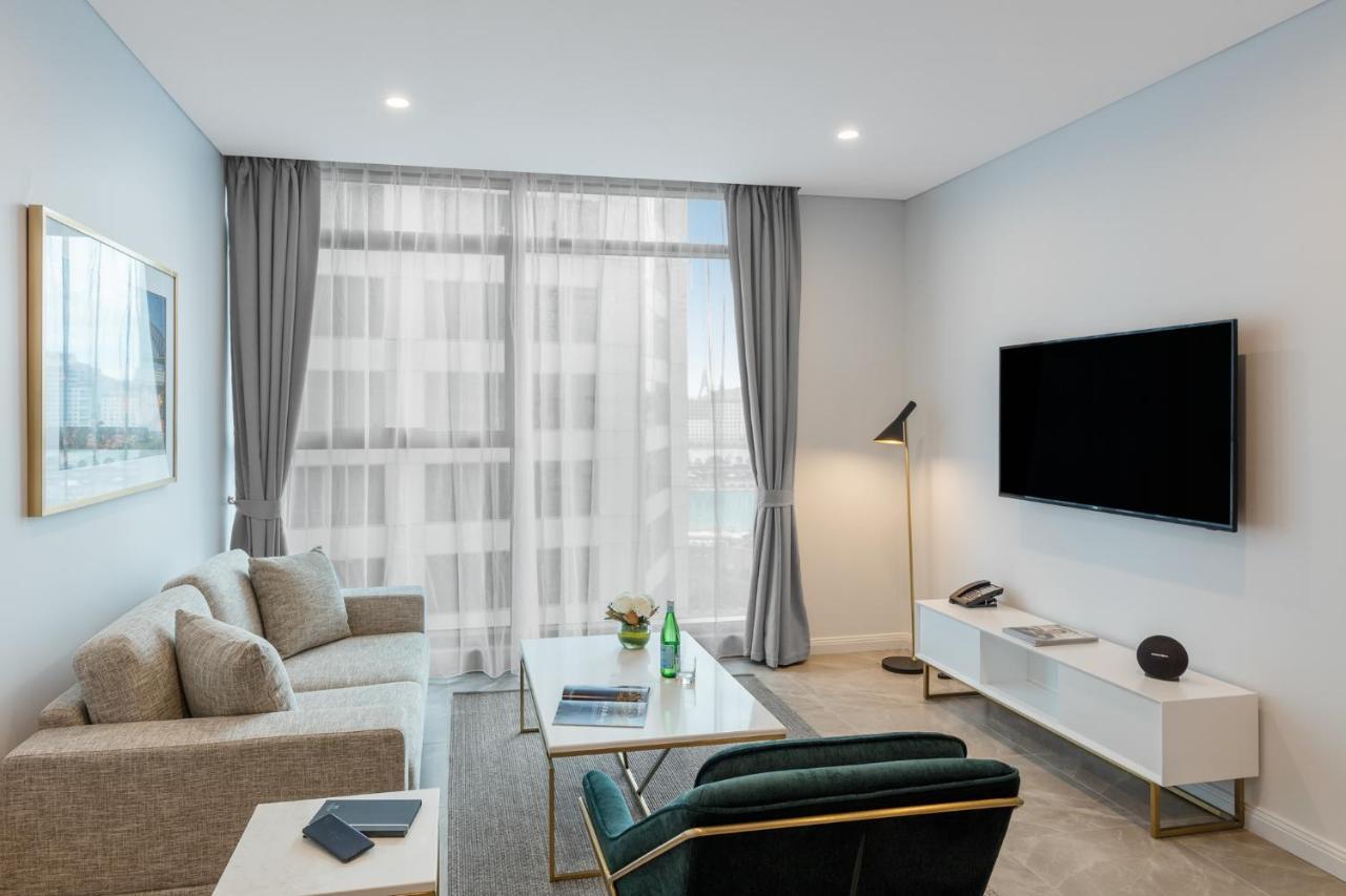 فندق أجنحة ميريتون سوسيكس ستريت سيدني المظهر الخارجي الصورة The photo shows a modern living room space with a light, airy ambiance. It features a comfortable beige sofa with decorative cushions, a sleek coffee table placed in front, and a dark green armchair. There is a large window covered with sheer curtain