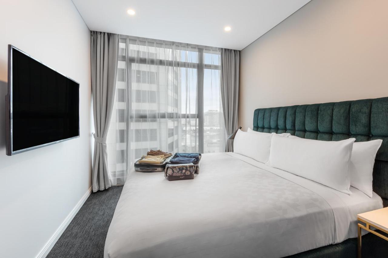 فندق أجنحة ميريتون سوسيكس ستريت سيدني المظهر الخارجي الصورة The photo shows a modern hotel bedroom. The room features a large bed with a green upholstered headboard and crisp white linens. There are two pillows on the bed and a few folded items of clothing neatly placed on it. A flat-screen TV is mounted on t