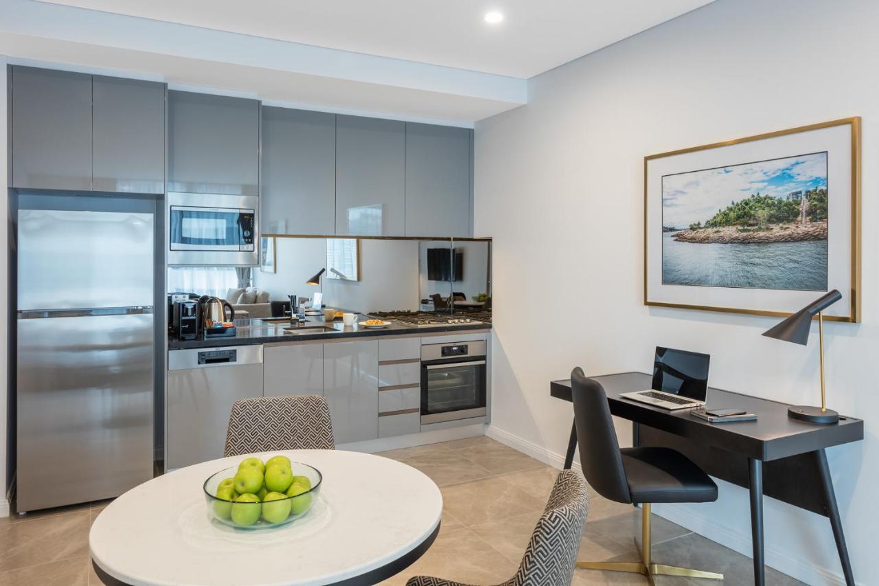 فندق أجنحة ميريتون سوسيكس ستريت سيدني المظهر الخارجي الصورة The photo shows a modern kitchen and dining area in an apartment. The kitchen features sleek gray cabinetry, a stainless steel fridge, and a microwave. There is a countertop with cooking appliances, including an oven and stovetop. A small round dinin