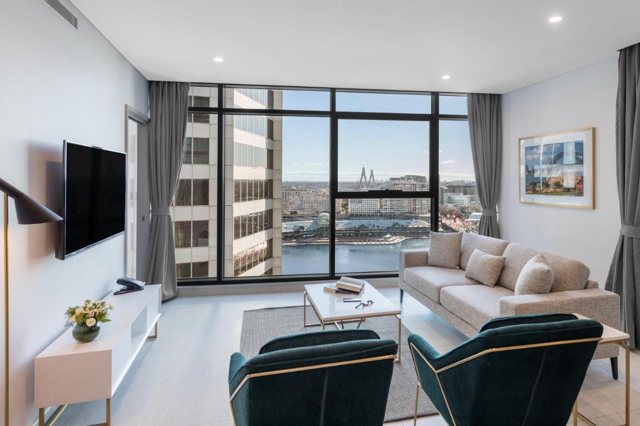 فندق أجنحة ميريتون سوسيكس ستريت سيدني المظهر الخارجي الصورة The photo shows a modern living room with large windows that offer a view of the cityscape and water. The interior features a light color palette with pale blue walls and soft lighting. There is a comfortable gray sofa and two green accent chairs, ar