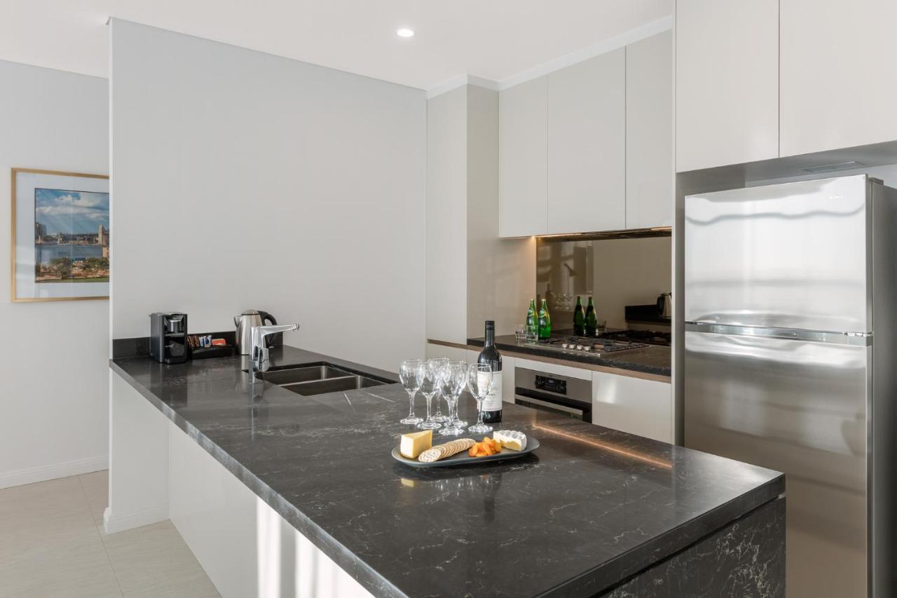 فندق أجنحة ميريتون سوسيكس ستريت سيدني المظهر الخارجي الصورة The photo shows a modern kitchen featuring a sleek design with a dark stone countertop. There is a built-in sink and possibly a cooktop. The kitchen cabinetry is light-colored, creating a contrast with the dark counter. On the countertop, there are w