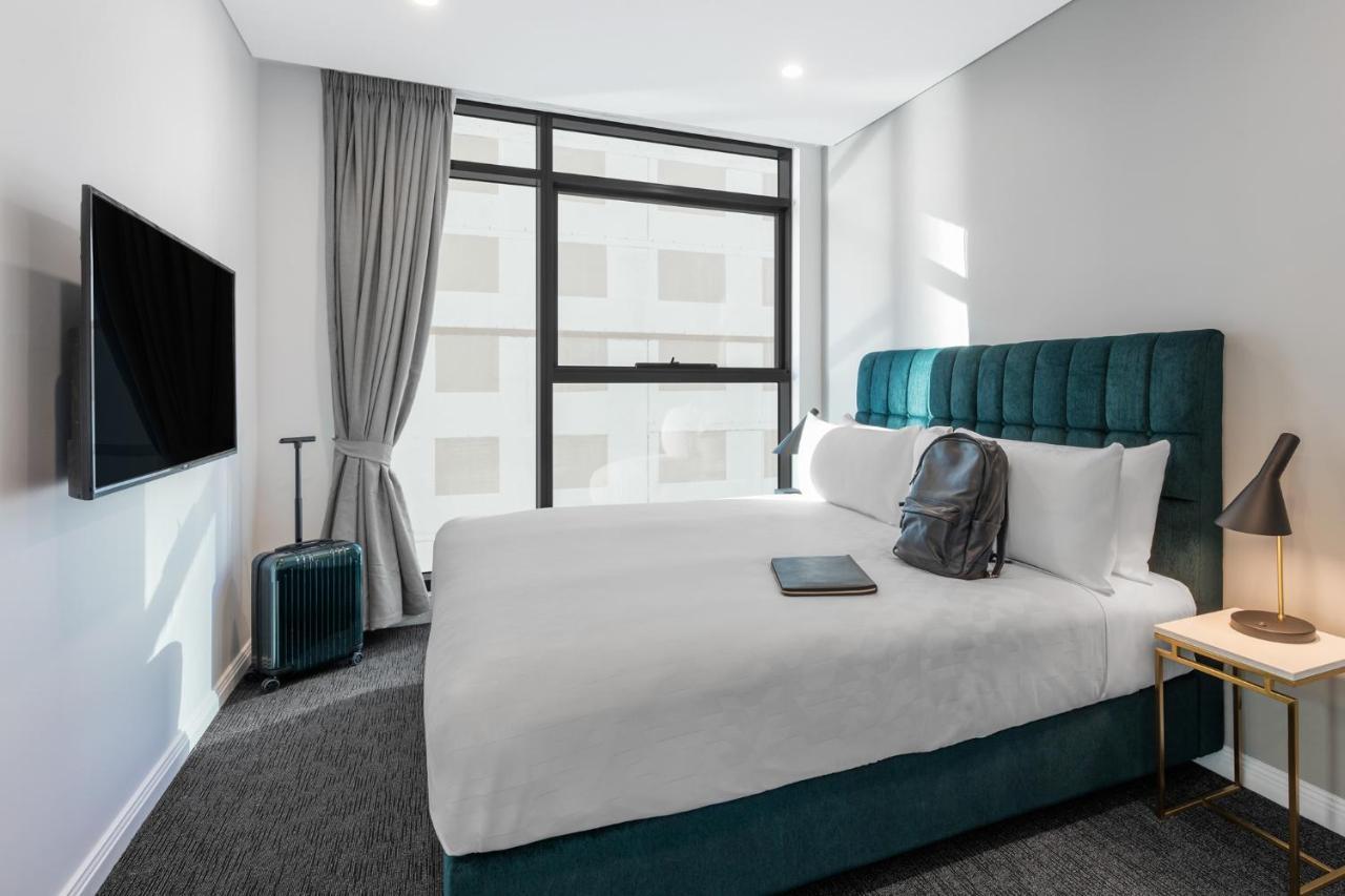 فندق أجنحة ميريتون سوسيكس ستريت سيدني المظهر الخارجي الصورة The photo shows a modern hotel room featuring a comfortable bed with white bedding and a teal-colored headboard. There is a neatly placed black backpack on the bed, along with a small notebook or tablet. A television is mounted on the wall opposite t