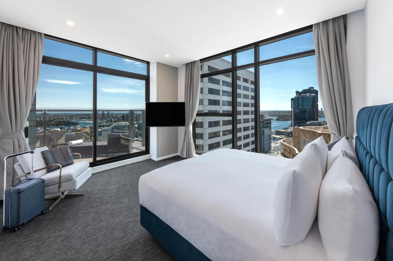فندق أجنحة ميريتون سوسيكس ستريت سيدني المظهر الخارجي الصورة The photo shows a modern hotel room featuring large windows that provide a panoramic view of the cityscape and waterfront. The room is furnished with a king-size bed dressed in white linens, a comfortable armchair, and a flat-screen TV mounted on the
