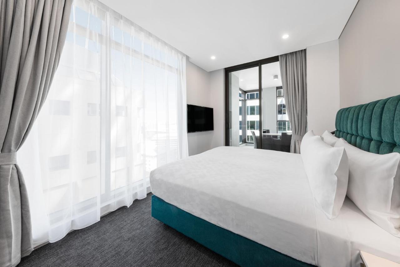 فندق أجنحة ميريتون سوسيكس ستريت سيدني المظهر الخارجي الصورة The photo shows a modern bedroom featuring a large bed with a white duvet and pillows. The headboard is turquoise, adding a pop of color to the neutral-toned room. There are sheer white curtains covering large windows that let in natural light, creat