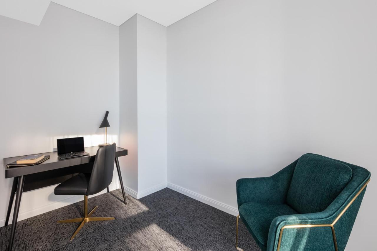 فندق أجنحة ميريتون سوسيكس ستريت سيدني المظهر الخارجي الصورة The photo depicts a minimalist interior space, likely a small office or study area. It features a simple, modern desk with a black lamp and a laptop or tablet on it. A sleek black chair accompanies the desk, while on the opposite side of the room, th