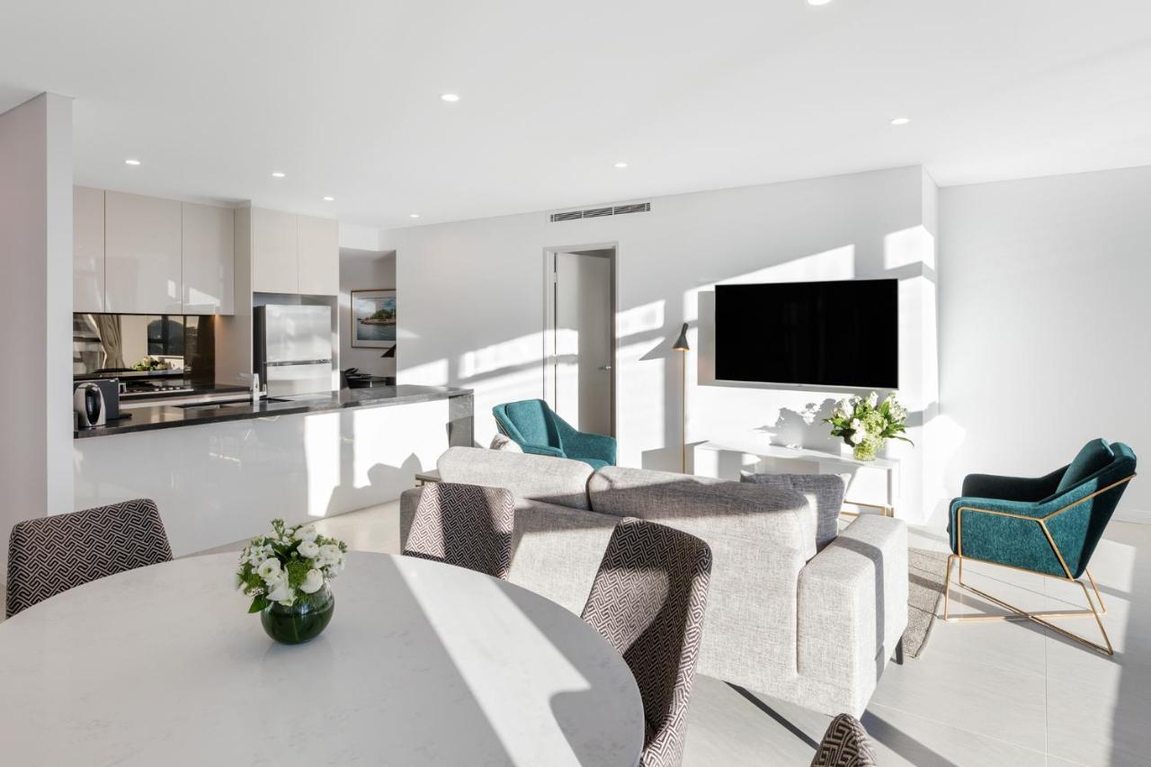 فندق أجنحة ميريتون سوسيكس ستريت سيدني المظهر الخارجي الصورة The photo shows a modern, bright living space that combines a kitchen and a living area. The kitchen features sleek cabinets and stainless steel appliances. In the foreground, there is a round dining table with a vase of flowers on it. Seating option