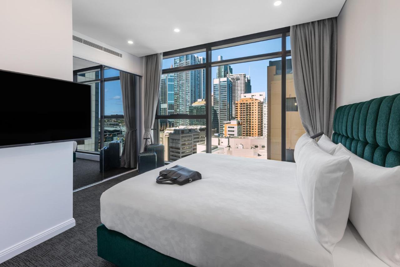 فندق أجنحة ميريتون سوسيكس ستريت سيدني المظهر الخارجي الصورة The photo shows a modern hotel room featuring a large bed with white linens and a green upholstered headboard. There is a sleek television mounted on the wall opposite the bed, and the room has large windows that provide a view of a cityscape, includ