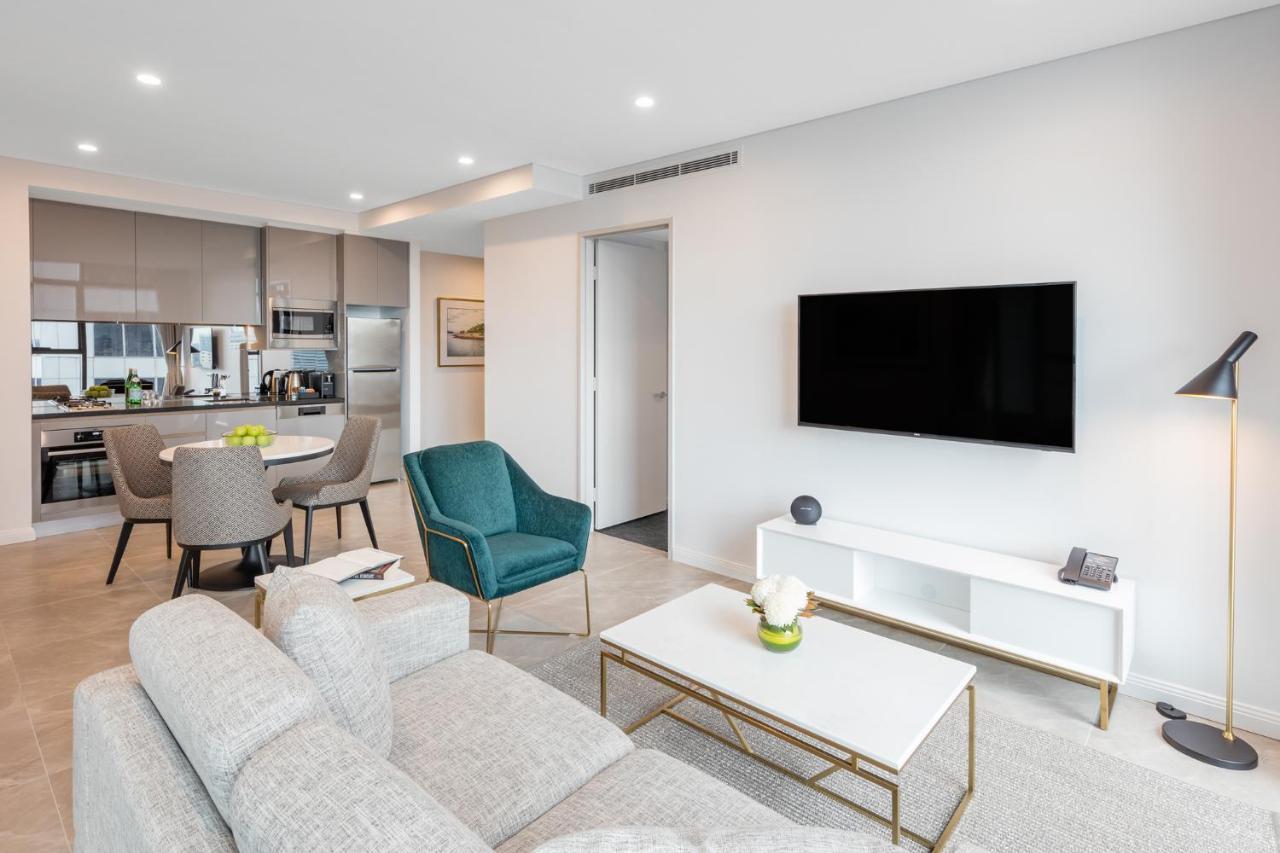 فندق أجنحة ميريتون سوسيكس ستريت سيدني المظهر الخارجي الصورة The photo shows a modern living room area. It features a light gray sofa and a coffee table with a decorative plant. There are two chairs, one in teal and a round one, positioned around the coffee table. A flat-screen TV is mounted on the wall, and t