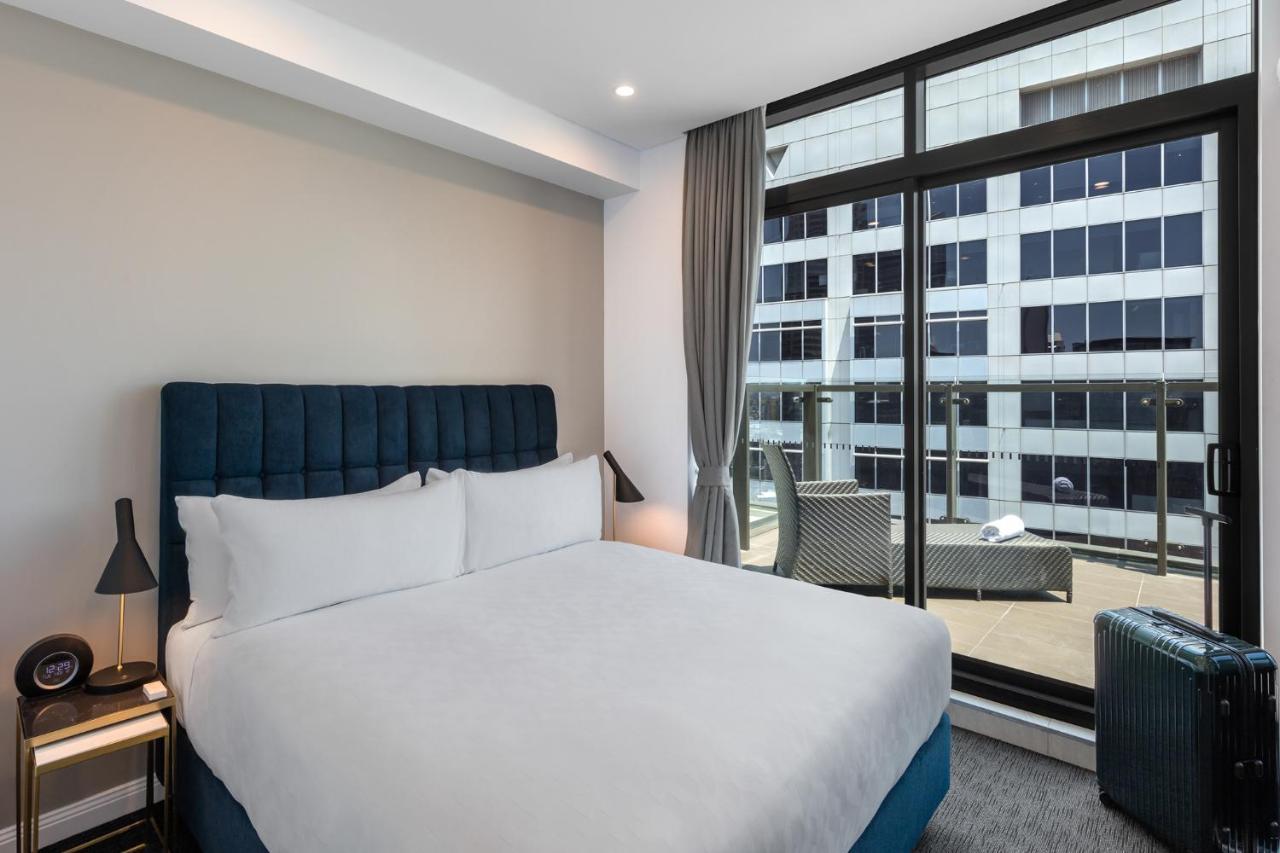 فندق أجنحة ميريتون سوسيكس ستريت سيدني المظهر الخارجي الصورة The photo shows a modern hotel room with a large bed featuring a blue upholstered headboard and white linens. Natural light enters through a large window, revealing a view of a building exterior, likely part of the hotel or surrounding area. The room