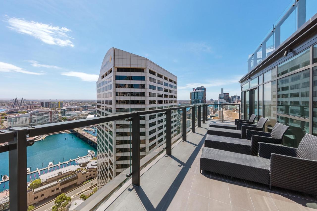 فندق أجنحة ميريتون سوسيكس ستريت سيدني المظهر الخارجي الصورة The photo shows a balcony or terrace with a panoramic view of a city skyline. The balcony features modern outdoor furniture, including sleek lounge chairs. In the background, you can see a waterfront area with buildings and a harbor, along with a cle