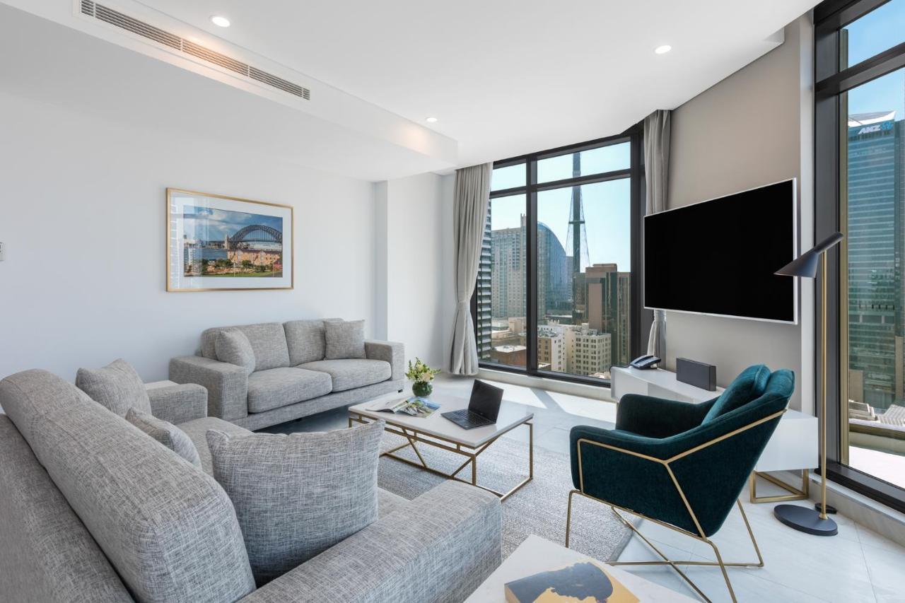 فندق أجنحة ميريتون سوسيكس ستريت سيدني المظهر الخارجي الصورة The photo shows a modern living room with large windows offering a view of a city skyline. The room features two gray sofas and a stylish green accent chair. A sleek coffee table is placed in front of the sofas, alongside a laptop. There’s a flat-scr