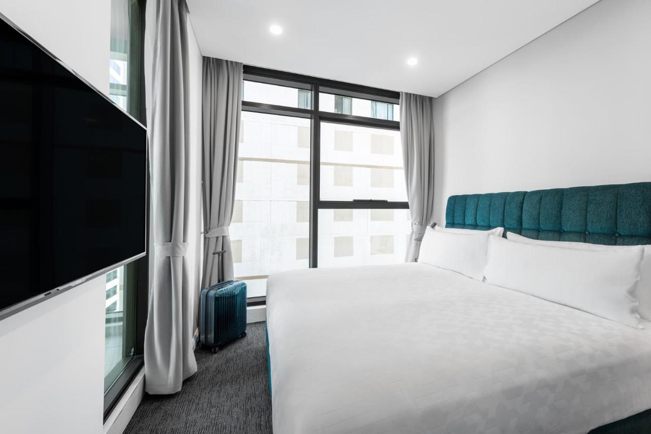 فندق أجنحة ميريتون سوسيكس ستريت سيدني المظهر الخارجي الصورة The photo shows a modern hotel room featuring a large bed with a light-colored duvet and a blue upholstered headboard. There are two pillows on the bed. A large window allows natural light to enter the room, and gray curtains frame the window. Next t