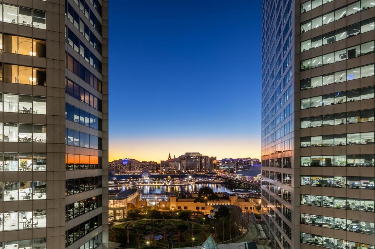 فندق أجنحة ميريتون سوسيكس ستريت سيدني المظهر الخارجي الصورة The photo shows a view between two tall buildings, likely office towers, during twilight. On one side, there are glass windows reflecting the evening sky that transitions from blue to orange near the horizon. In the background, a cityscape is visible