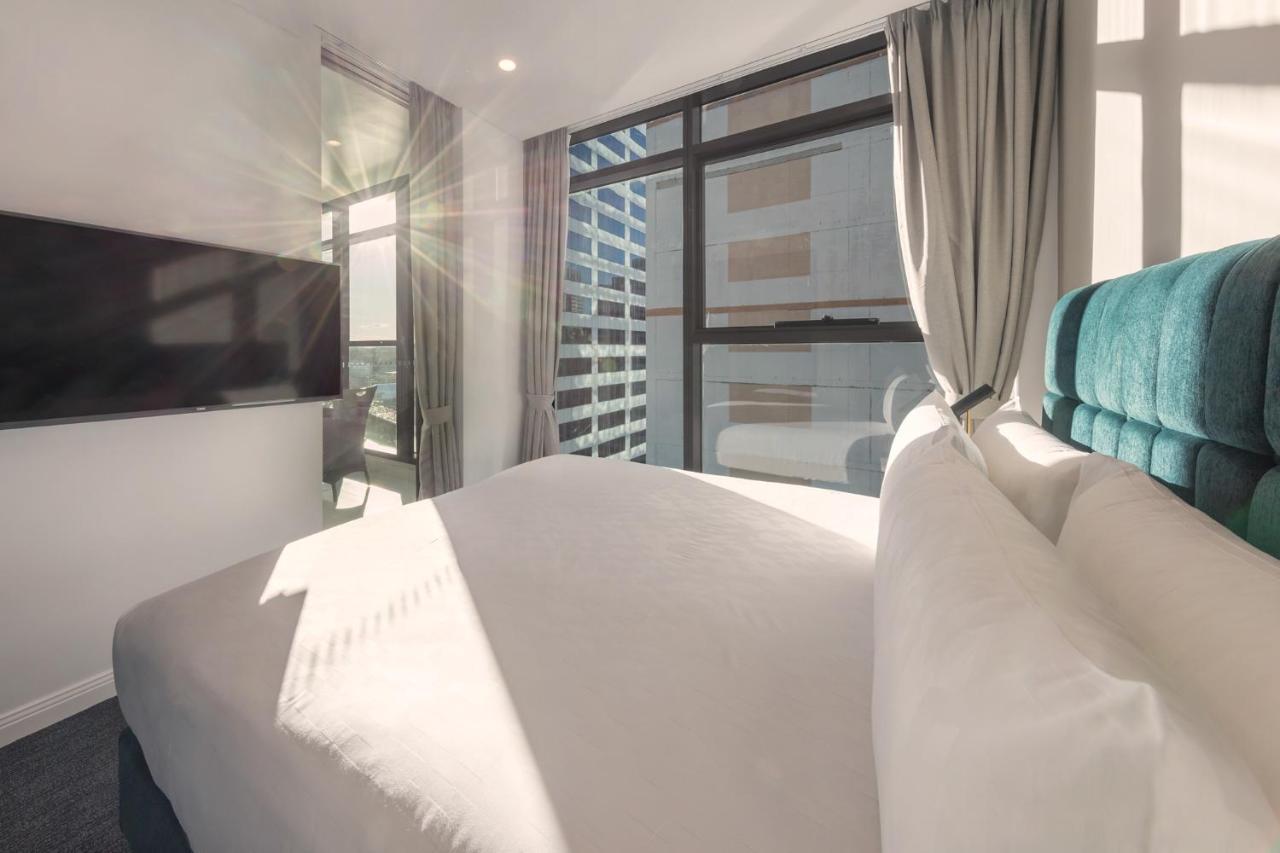 فندق أجنحة ميريتون سوسيكس ستريت سيدني المظهر الخارجي الصورة The photo shows a modern hotel room with a neatly made bed featuring white bedding and a plush, tufted headboard in a shade of blue. Natural light is streaming through large windows, which are covered partially by light gray curtains. A flat-screen t