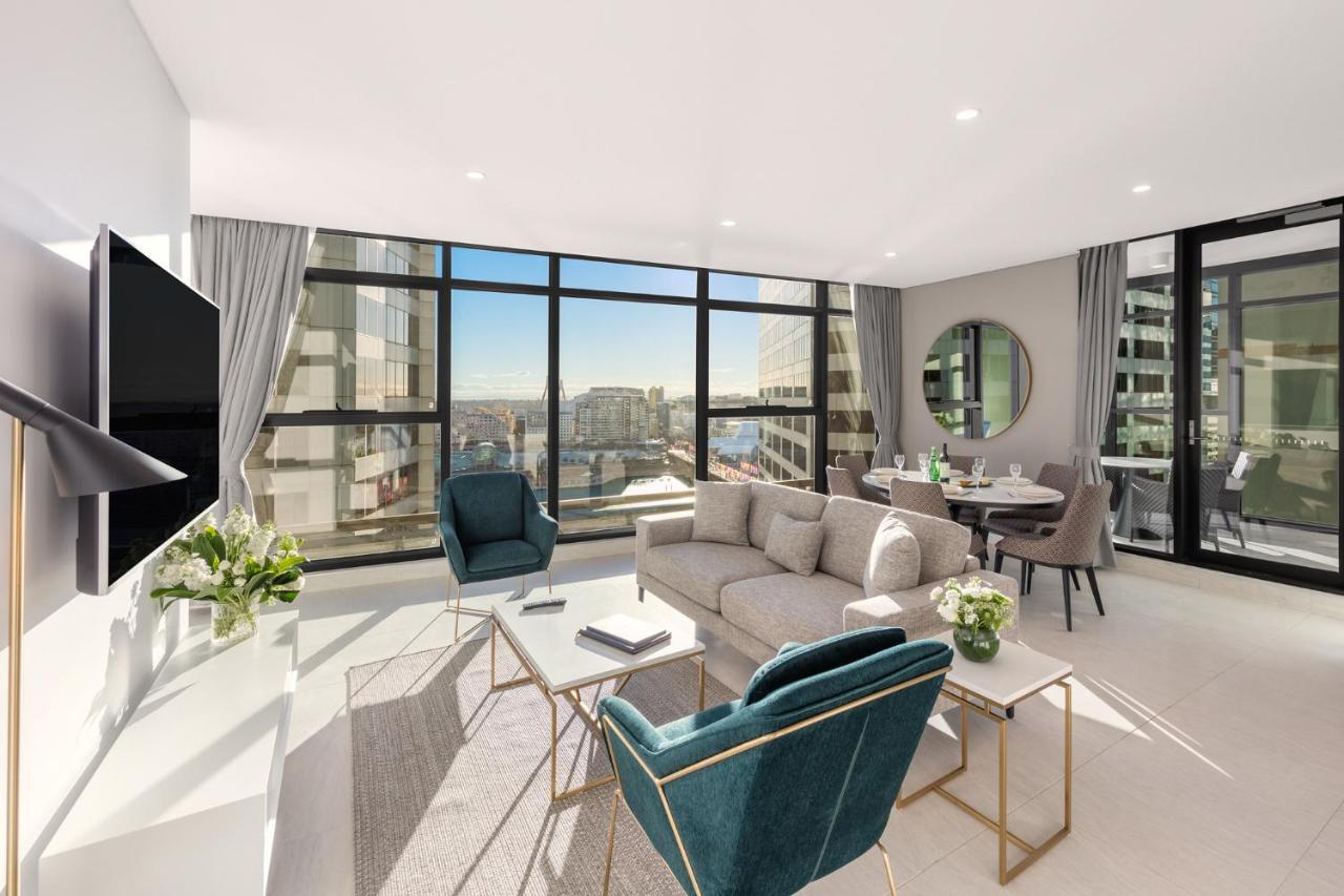 فندق أجنحة ميريتون سوسيكس ستريت سيدني المظهر الخارجي الصورة The photo shows a modern and stylish living space. It features a spacious room with large windows that provide a bright view of the cityscape. The furniture includes a light gray sofa, two teal chairs, and a coffee table. There is a flat-screen TV mo