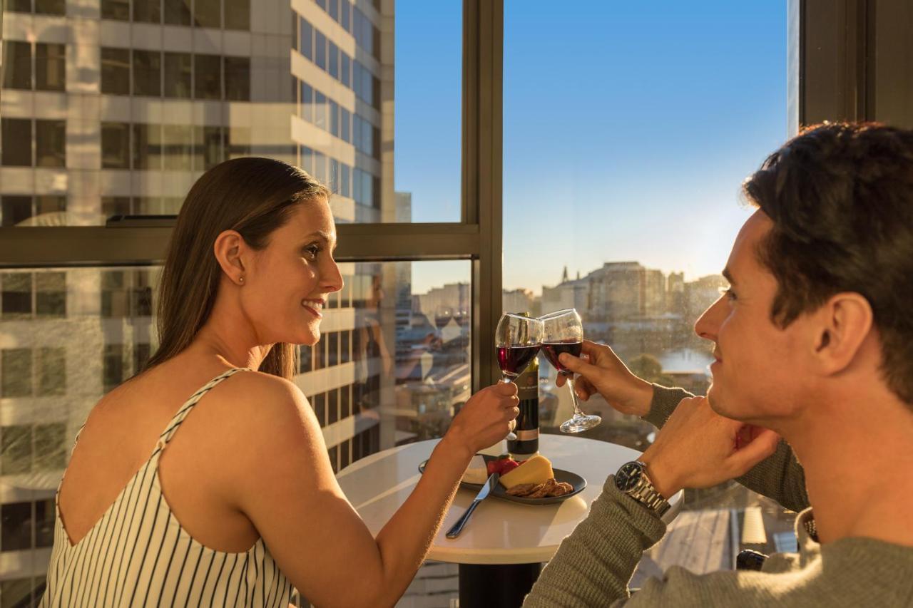 فندق أجنحة ميريتون سوسيكس ستريت سيدني المظهر الخارجي الصورة The photo depicts a couple enjoying a drink together in a modern setting. They are sitting at a small table near a large window, which provides a view of a cityscape and a sunset. Both individuals are smiling and engaged in a cheerful toast with glas