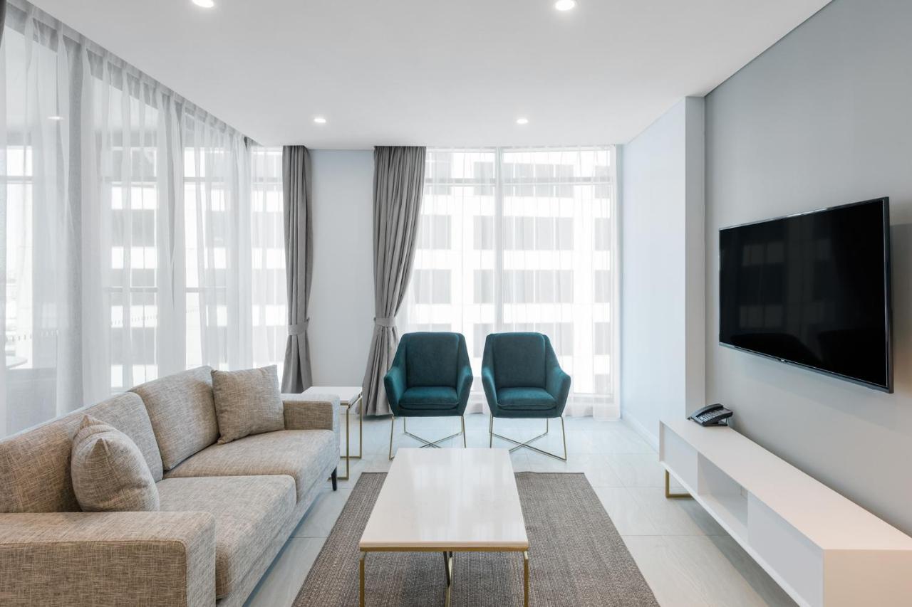 فندق أجنحة ميريتون سوسيكس ستريت سيدني المظهر الخارجي الصورة The photo shows a modern living room equipped with a light gray sofa, two teal chairs, and a coffee table in the center. The room features large windows with sheer curtains, allowing natural light to illuminate the space. There is a white media conso