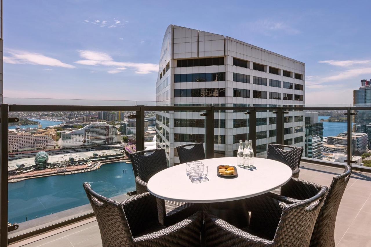 فندق أجنحة ميريتون سوسيكس ستريت سيدني المظهر الخارجي الصورة The photo shows a balcony or terrace with a round table and several chairs arranged around it. The table is set with some drinks and a plate, possibly containing food. In the background, there is a view of a waterway and several buildings, including 