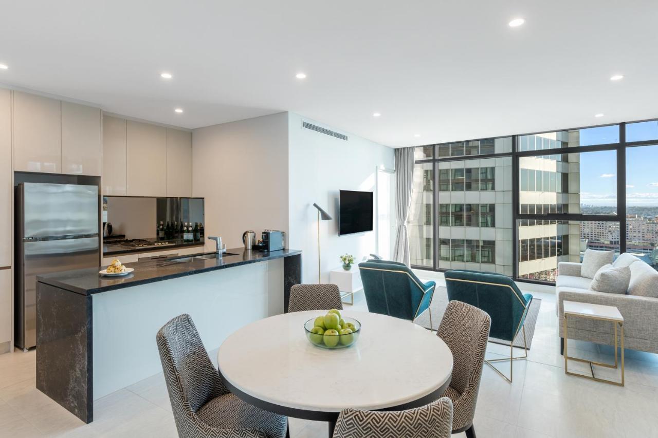 فندق أجنحة ميريتون سوسيكس ستريت سيدني المظهر الخارجي الصورة The photo shows a modern and stylish interior space, likely a living room and kitchen area. The kitchen features sleek cabinetry and a countertop, with appliances visible, including a coffee maker. There is a round dining table with gray chairs, set 