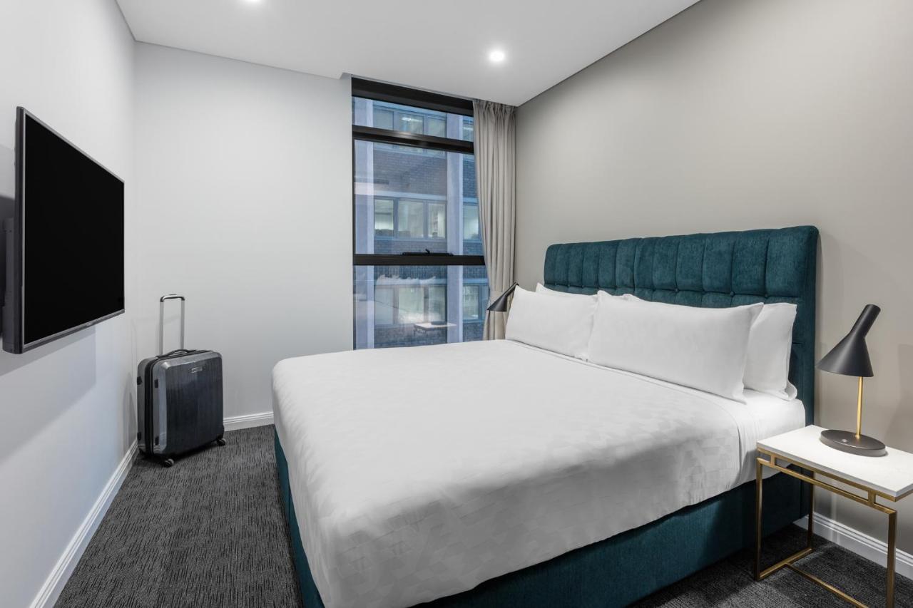 فندق أجنحة ميريتون سوسيكس ستريت سيدني المظهر الخارجي الصورة The photo shows a modern hotel room with a clean and minimalist design. There is a large, neatly made bed with white bedding and a teal-colored headboard. On one side of the bed, there is a small nightstand with a lamp. A sleek television is mounted 