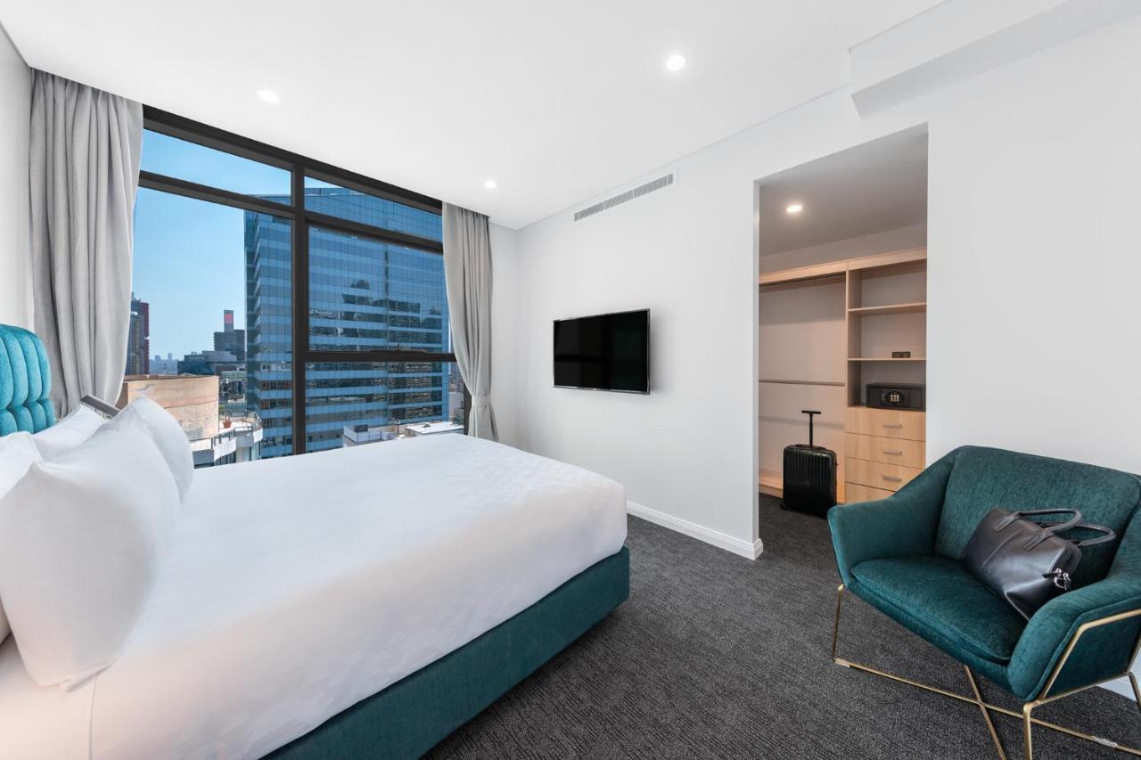 فندق أجنحة ميريتون سوسيكس ستريت سيدني المظهر الخارجي الصورة The photo shows a modern hotel room. It features a large window that allows natural light to fill the space, offering a view of the city skyline. The room includes a neatly made bed with a white duvet and a teal-colored base. There is a small green a