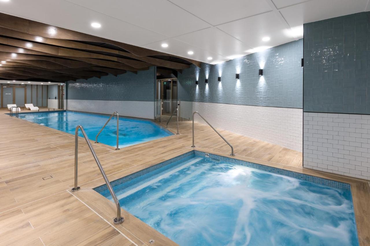 فندق أجنحة ميريتون سوسيكس ستريت سيدني المظهر الخارجي الصورة The photo shows a modern indoor spa area. There are two pools: one is larger and has a clear blue water, while the other is smaller, possibly a hot tub, with jets creating bubbles. The walls are a mix of blue tiles and white tiles, and the floor is m