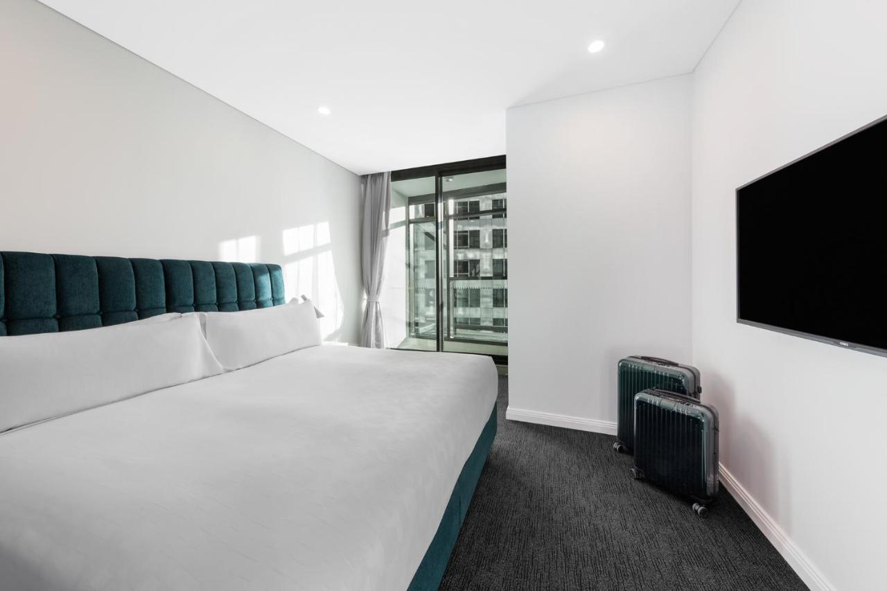 فندق أجنحة ميريتون سوسيكس ستريت سيدني المظهر الخارجي الصورة The photo shows a modern hotel room featuring a large bed with a headboard in a teal color. The bedding is white and appears neatly arranged. There is a large window allowing natural light to enter, offering a view of an outdoor area. A flat-screen T