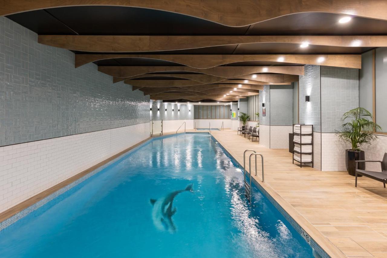 فندق أجنحة ميريتون سوسيكس ستريت سيدني المظهر الخارجي الصورة The photo shows an indoor swimming pool. The pool features clear blue water, and there is a dolphin swimming in it. The surrounding area has a modern design with curved wooden beams on the ceiling, and the walls are tiled in light colors. There are a