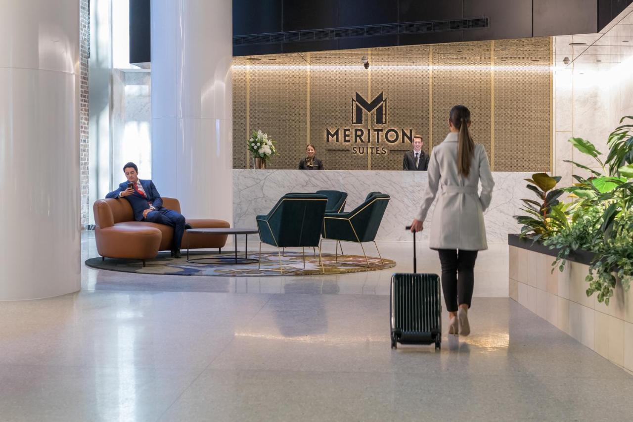 فندق أجنحة ميريتون سوسيكس ستريت سيدني المظهر الخارجي الصورة The photo depicts a modern hotel lobby. In the foreground, a woman is walking towards the reception desk, pulling a suitcase behind her. She is dressed in a light-colored coat. To her left, there’s a comfortable seating area with a person sitting on 