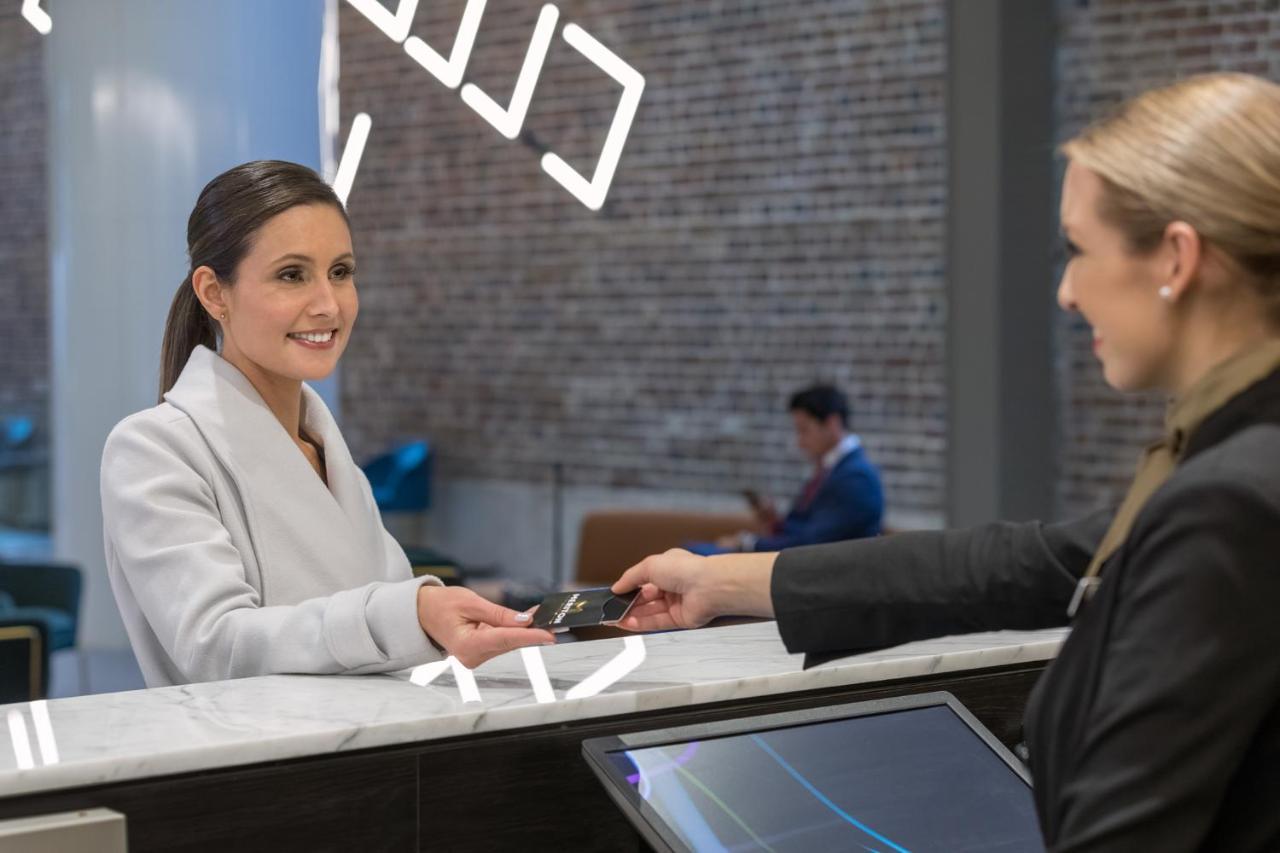 فندق أجنحة ميريتون سوسيكس ستريت سيدني المظهر الخارجي الصورة The image depicts a scene at a hotel reception desk. A woman with long hair, dressed in a white outfit, is smiling as she hands her card to a receptionist, who is in a black uniform. The reception area features a modern design, and there is a person 
