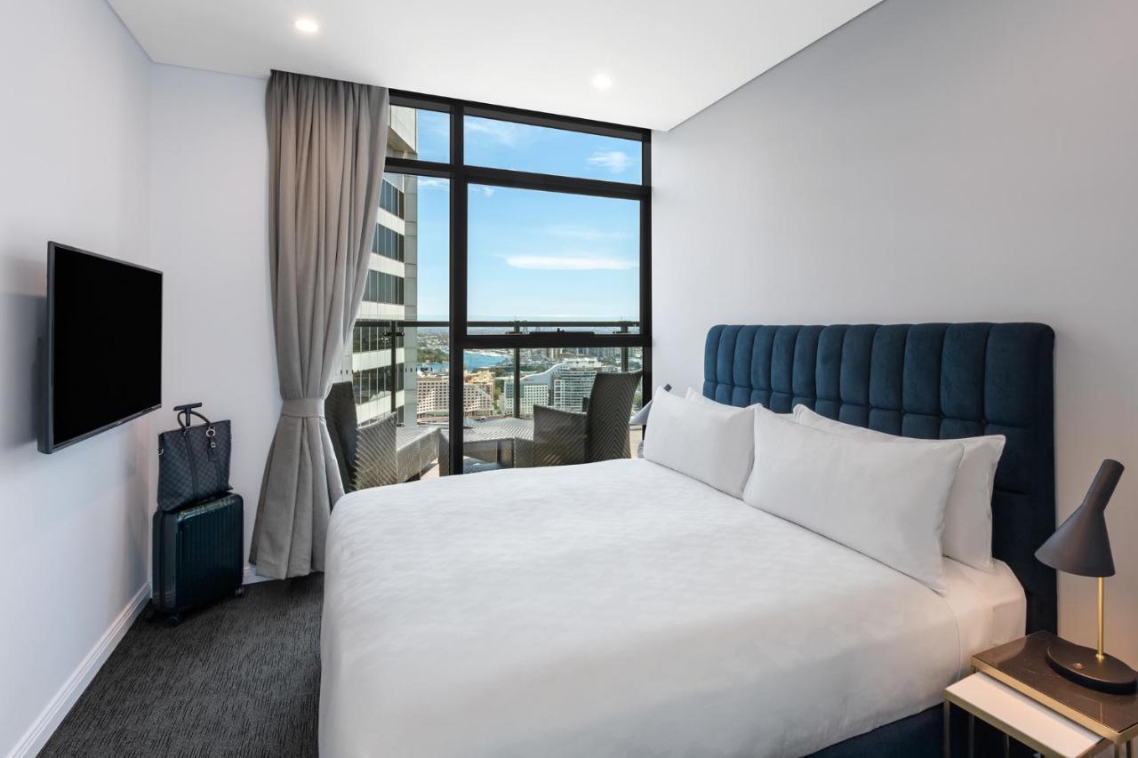 فندق أجنحة ميريتون سوسيكس ستريت سيدني المظهر الخارجي الصورة The photo shows a modern hotel room featuring a double bed with white linens and a tufted blue headboard. There is a small bedside table next to the bed. A large window allows natural light to fill the room, offering a view of the city outside, inclu