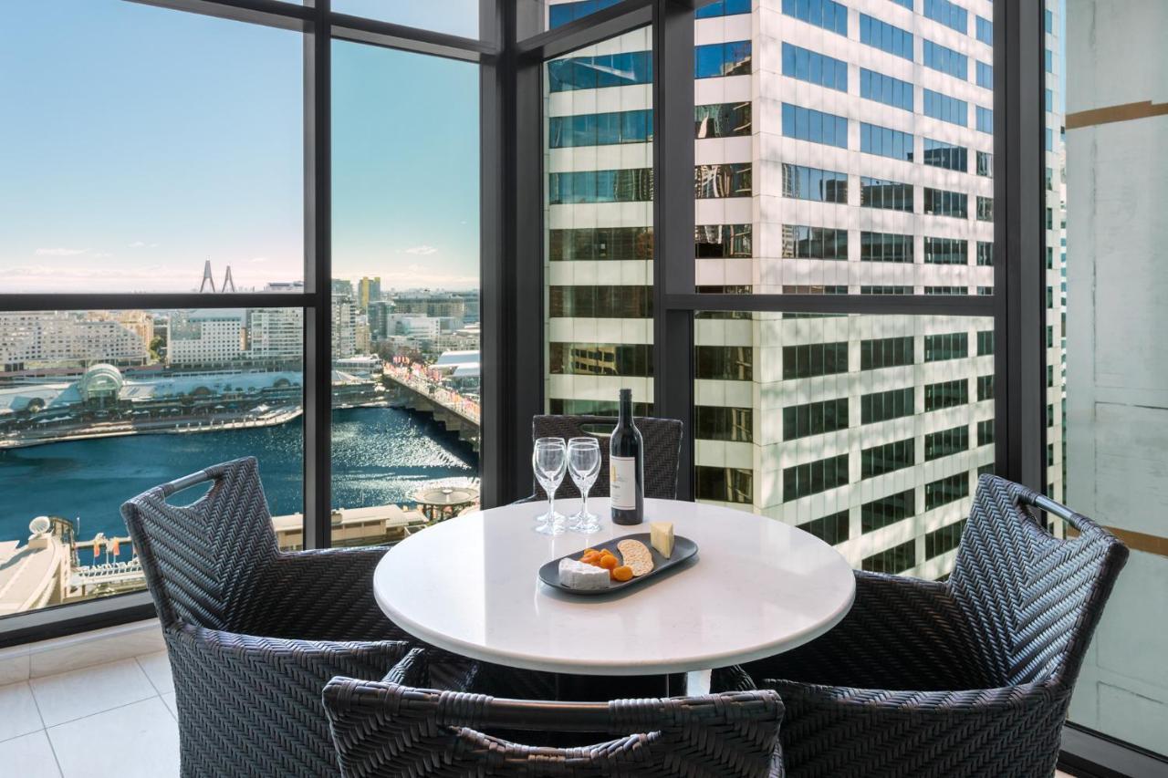 فندق أجنحة ميريتون سوسيكس ستريت سيدني المظهر الخارجي الصورة The photo shows a modern dining area with a round table and four woven chairs. On the table, there are two wine glasses, a bottle of wine, and a plate with food items. The large windows provide a view of a cityscape featuring tall buildings and a bod