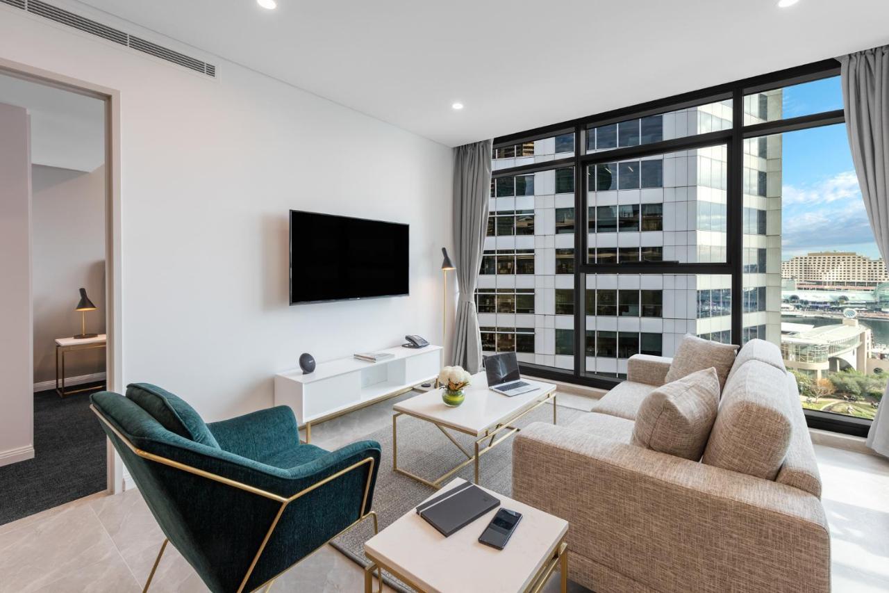 فندق أجنحة ميريتون سوسيكس ستريت سيدني المظهر الخارجي الصورة The photo shows a modern living room with large windows that let in natural light, providing a view of a cityscape outside. The interior features a comfortable light-colored sofa and a stylish dark green armchair. A sleek TV is mounted on the wall op