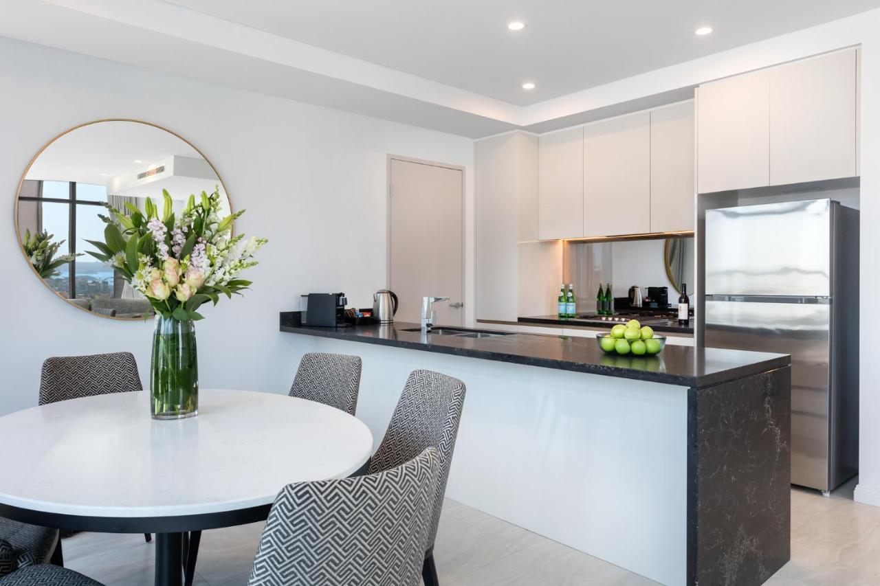 فندق أجنحة ميريتون سوسيكس ستريت سيدني المظهر الخارجي الصورة The photo shows a modern kitchen and dining area. In the foreground, there's a circular dining table surrounded by four stylish chairs. A large vase filled with fresh flowers is placed on the table, adding a touch of color and elegance. 

In the kitc