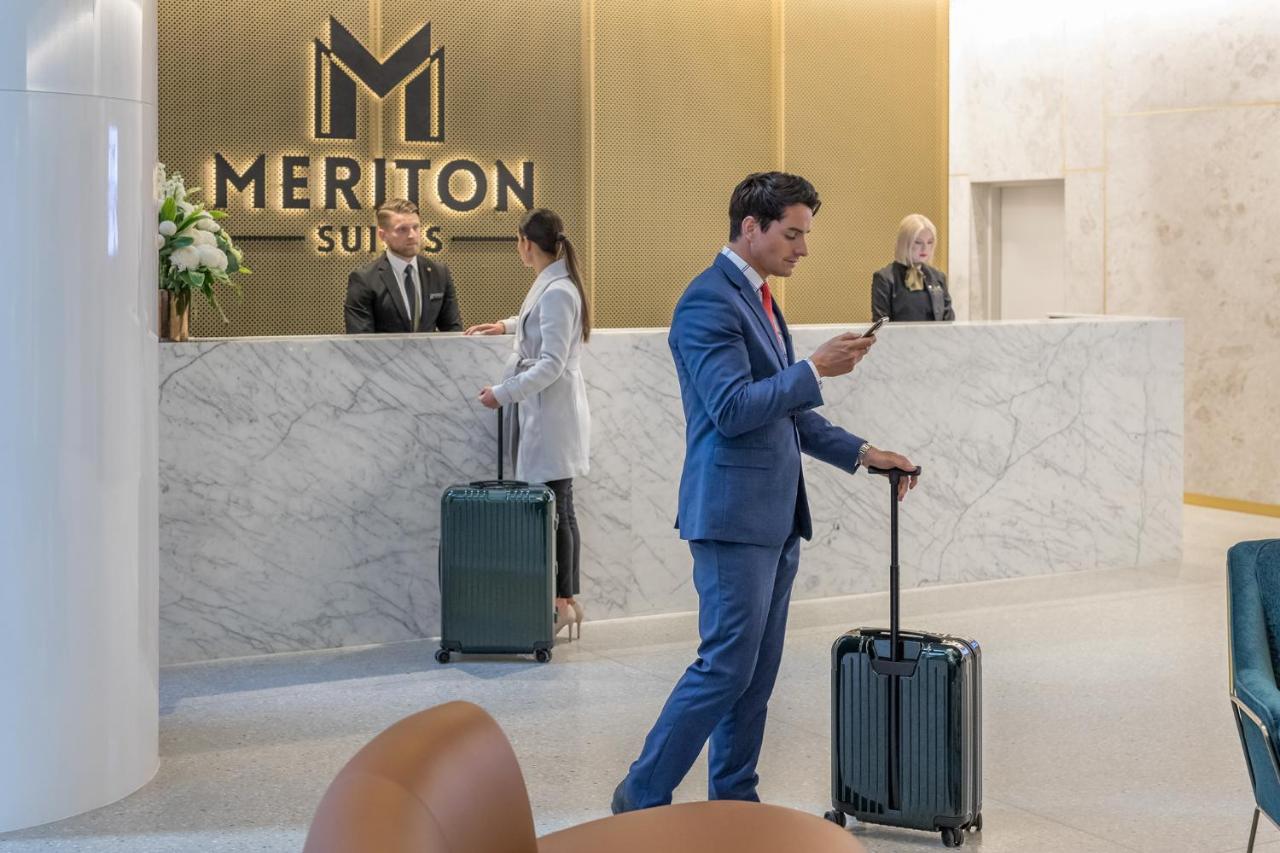 فندق أجنحة ميريتون سوسيكس ستريت سيدني المظهر الخارجي الصورة The photo shows the interior of a hotel reception area, specifically at Meriton Suites. In the foreground, a man in a blue suit is standing with a suitcase, looking at his phone. Behind him, there are two staff members at the reception desk, one of w