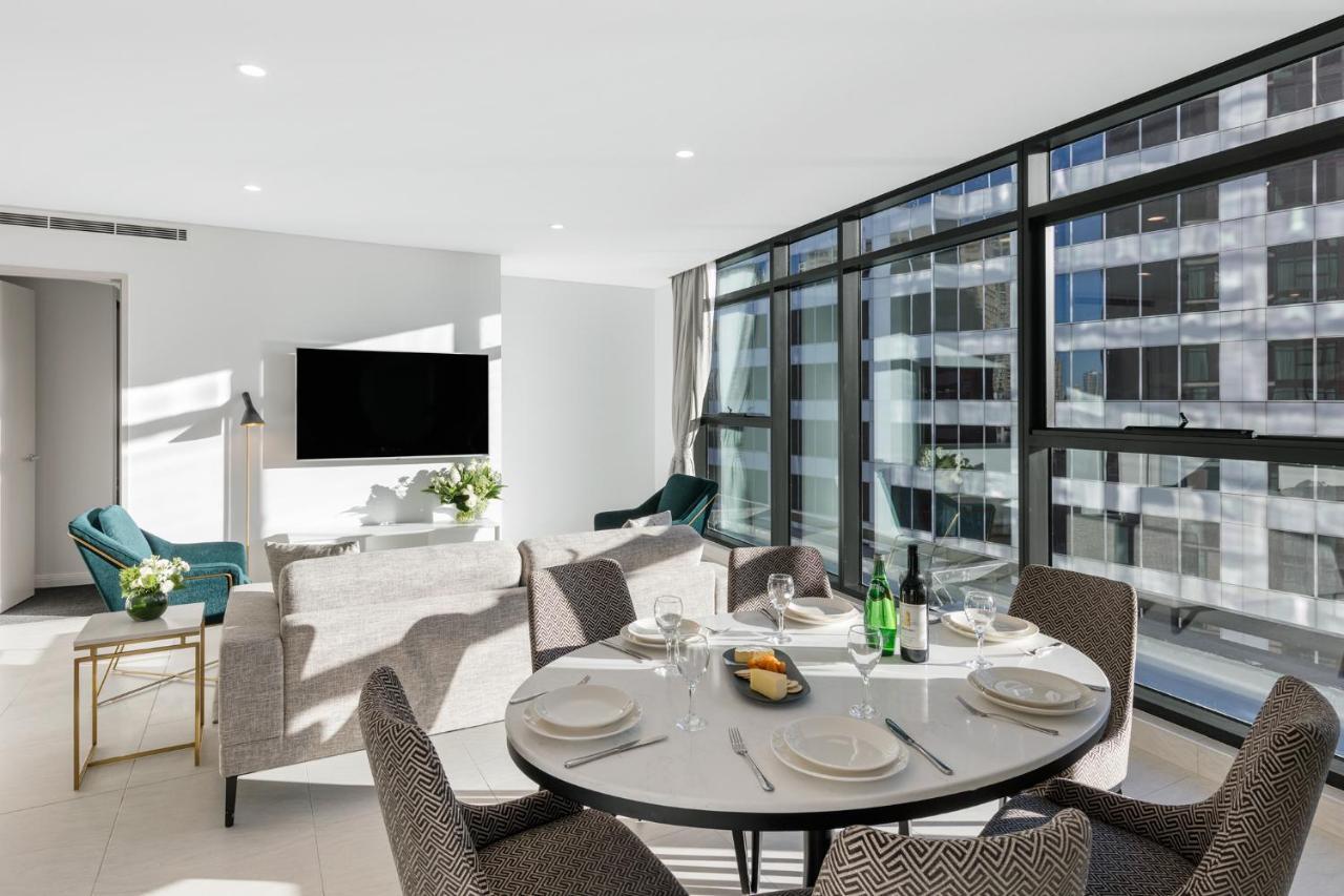 فندق أجنحة ميريتون سوسيكس ستريت سيدني المظهر الخارجي الصورة The photo shows a modern and stylish living room and dining area. In the foreground, there is a round dining table set for a meal with plates, cutlery, and drinks. The seating arrangement includes comfortable chairs around the table. 

In the backgro