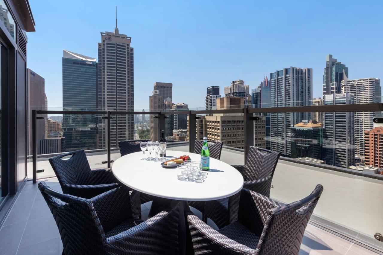 فندق أجنحة ميريتون سوسيكس ستريت سيدني المظهر الخارجي الصورة The photo shows a stylish outdoor dining area on a balcony with a round white table surrounded by black wicker chairs. On the table, there are several glasses, a bottle of sparkling water, and a plate of snacks. In the background, there is a panorami