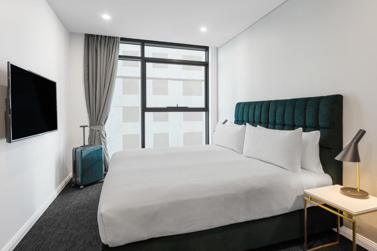 فندق أجنحة ميريتون سوسيكس ستريت سيدني المظهر الخارجي الصورة The photo shows a modern hotel room. It features a large bed with a dark green tufted headboard and multiple white pillows. There's a nightstand with a lamp beside the bed. A sleek television is mounted on the wall opposite the bed, and a suitcase is