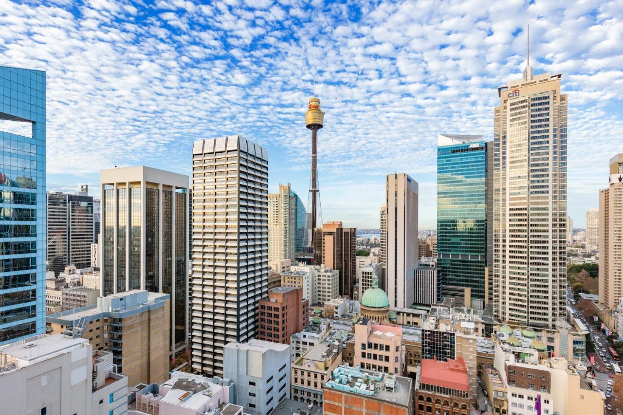 فندق أجنحة ميريتون سوسيكس ستريت سيدني المظهر الخارجي الصورة The photo shows a panoramic view of a city skyline dominated by tall skyscrapers. In the center, there is a notable tower with a distinctive golden structure at its top, which is likely a landmark of the city. The buildings have a mix of modern glass