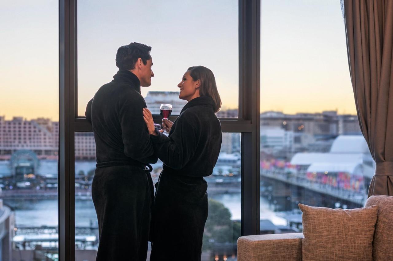 فندق أجنحة ميريتون سوسيكس ستريت سيدني المظهر الخارجي الصورة The photo depicts a couple standing closely together by a large window. They are wearing dark bathrobes and holding drinks, gazing into each other’s eyes. The background features a scenic view, likely of a cityscape at sunset, with soft light illumin