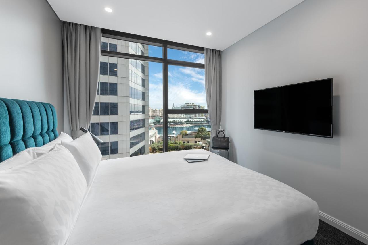 فندق أجنحة ميريتون سوسيكس ستريت سيدني المظهر الخارجي الصورة The photo shows a modern hotel room with a large bed featuring a white comforter and pillows. There is a blue headboard that adds a pop of color to the decor. A large window provides a view of the outside, which includes buildings and a glimpse of wa