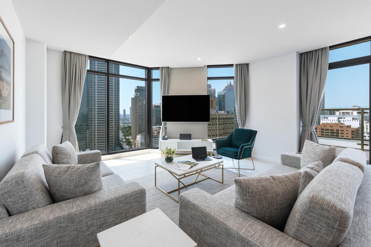 فندق أجنحة ميريتون سوسيكس ستريت سيدني المظهر الخارجي الصورة The photo showcases a modern living room with large windows offering a view of a city skyline. The room features a light color palette with gray sofas arranged around a coffee table. A flat-screen TV is mounted on the wall, and there is a green armch