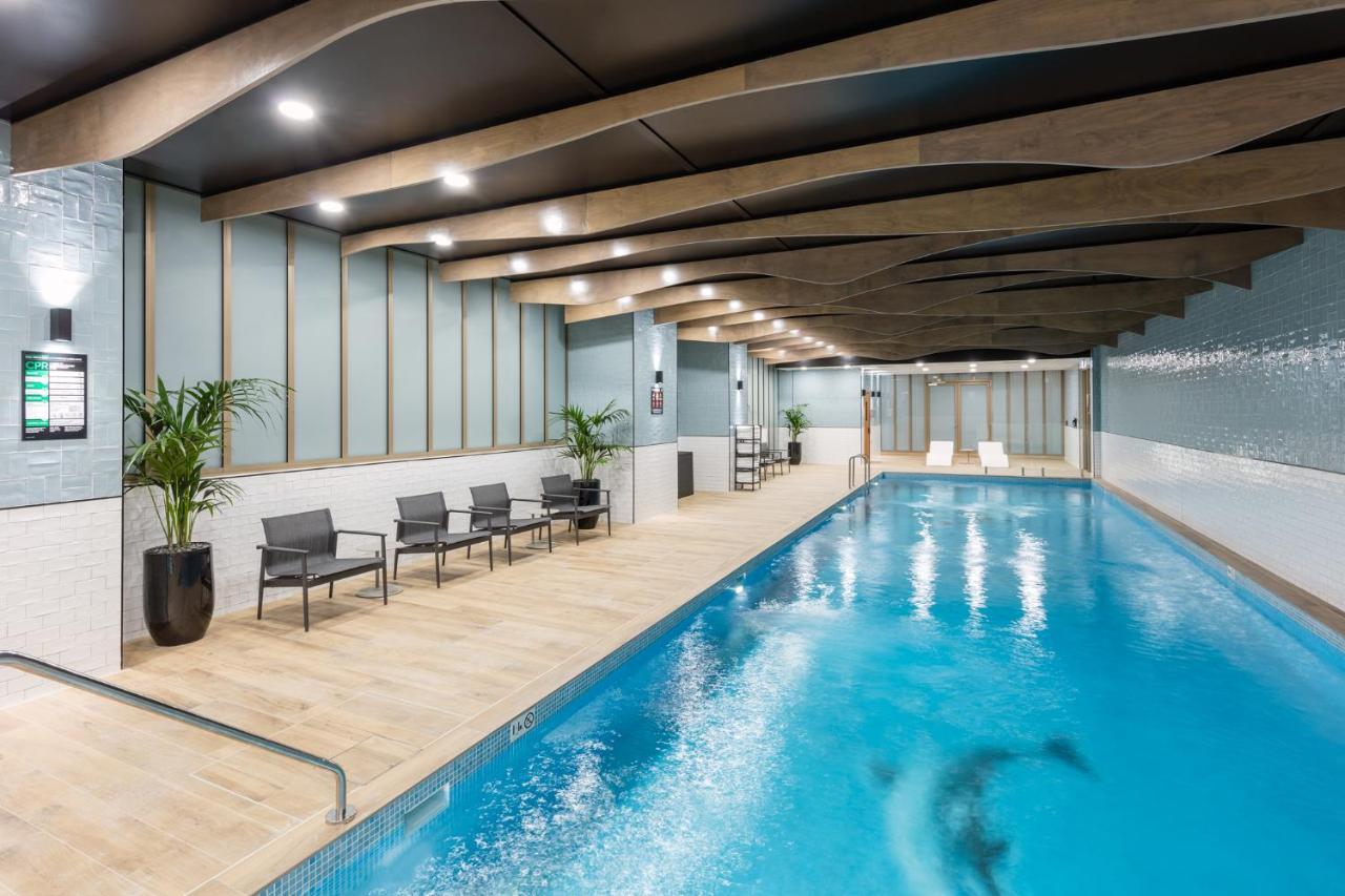 فندق أجنحة ميريتون سوسيكس ستريت سيدني المظهر الخارجي الصورة The photo shows an indoor swimming pool area. The pool has clear blue water and is flanked by a wooden deck. There are several lounge chairs arranged along one side of the pool. The ceiling features a modern design with wooden beams and recessed ligh