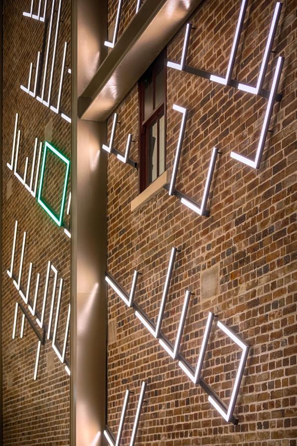 فندق أجنحة ميريتون سوسيكس ستريت سيدني المظهر الخارجي الصورة The photo shows a brick wall featuring a design made up of linear light fixtures. These light strips create geometric shapes and patterns, with some sections illuminated in white and one section highlighted in green. The combination of the brick text