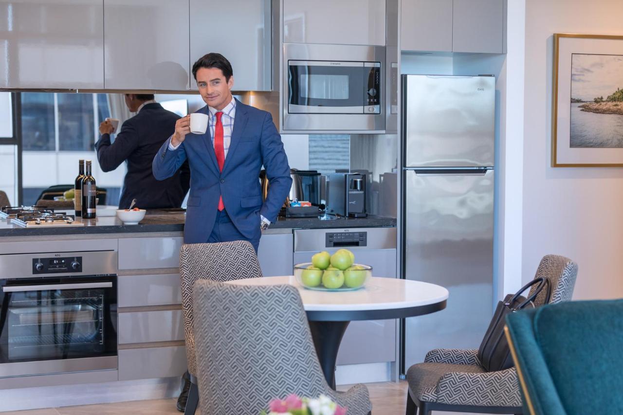 فندق أجنحة ميريتون سوسيكس ستريت سيدني المظهر الخارجي الصورة The photo depicts a well-dressed man in a suit standing in a modern kitchen. He is holding a cup, possibly of coffee, and appears to be looking towards the camera. The kitchen features contemporary appliances, including a microwave and refrigerator. 