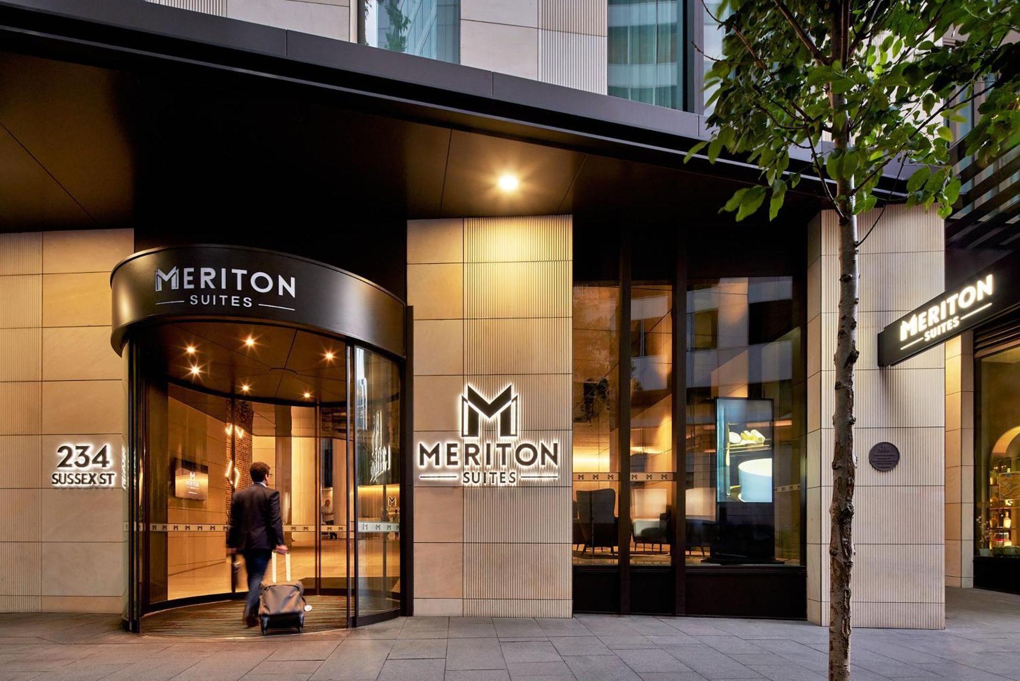 فندق أجنحة ميريتون سوسيكس ستريت سيدني المظهر الخارجي الصورة The photo shows the entrance of a hotel or suite establishment named "Meriton Suites." The entrance features a revolving door and a large, illuminated sign with the name "MERITON SUITES." The facade is modern and sleek, with a combination of glass an