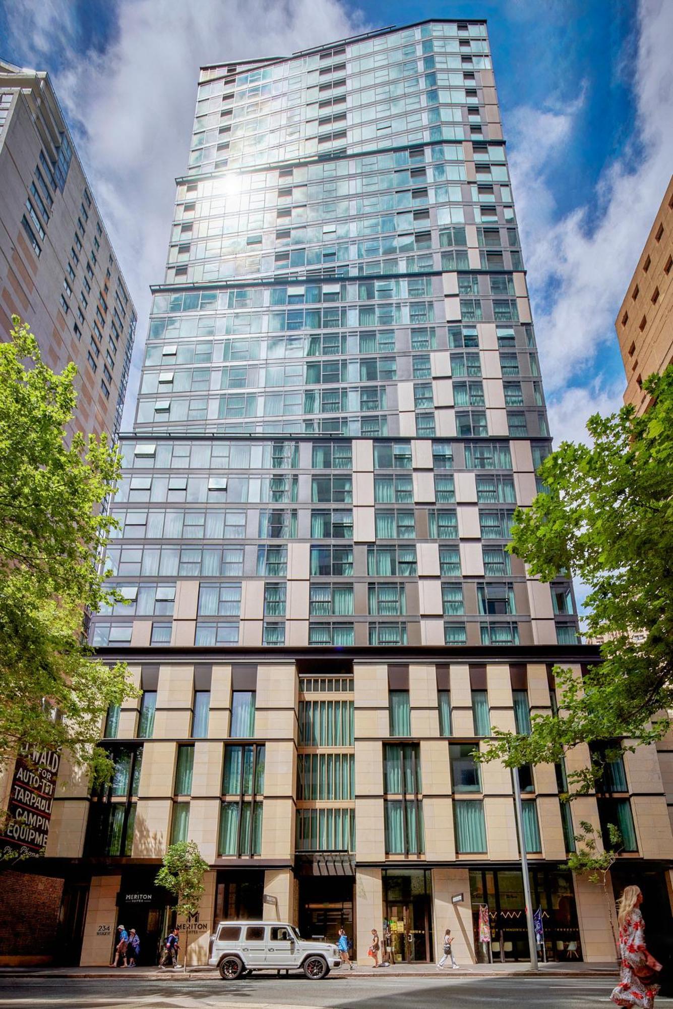 فندق أجنحة ميريتون سوسيكس ستريت سيدني المظهر الخارجي الصورة The photo shows a tall modern building with a glass facade and various architectural design elements. The structure has multiple stories, with large windows that reflect the surrounding environment. At the base, there are sections that appear to have